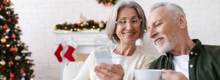 smiling and mature woman in glasses holding smartphone near husband with cup of tea, banner