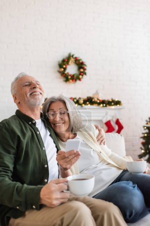 Téléchargez les photos : Femme gaie et d'âge moyen dans des lunettes tenant smartphone près mari heureux avec tasse de thé - en image libre de droit