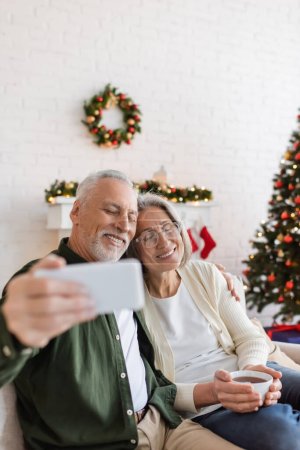 hombre de mediana edad tomando selfie con esposa feliz cerca del árbol de Navidad en casa