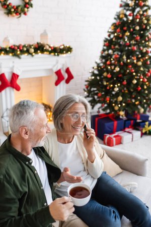 Téléchargez les photos : Homme d'âge moyen tenant tasse de thé et regardant femme parler sur smartphone près de l'arbre de Noël - en image libre de droit