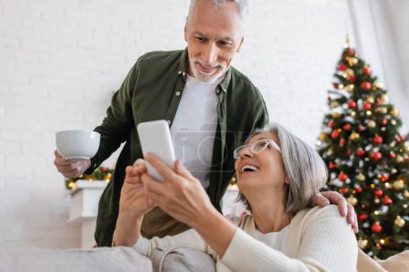 Reifer Mann mit Tasse Tee und Blick auf Smartphone neben Frau und verschwommenem Weihnachtsbaum