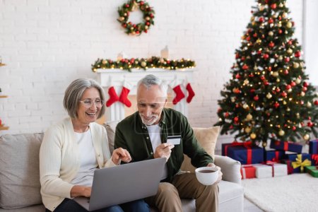 happy mature man holding credit card near wife with laptop while having online shopping during christmas 