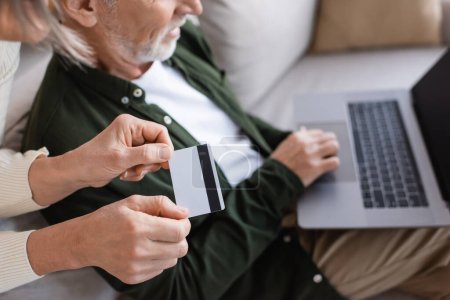 cropped view of smiling mature man using laptop near wife with credit card 