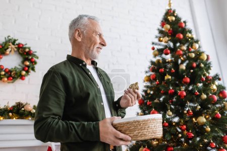 smiling middle aged man with beard holding wicker basket and baubles near christmas tree 