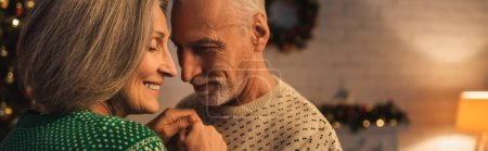 cheerful bearded man in festive sweater hugging smiling mature wife on christmas evening, banner