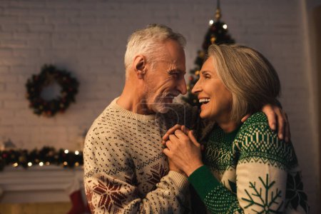 Téléchargez les photos : Gai mature homme en chandail festif étreignant femme joyeuse le soir de Noël - en image libre de droit