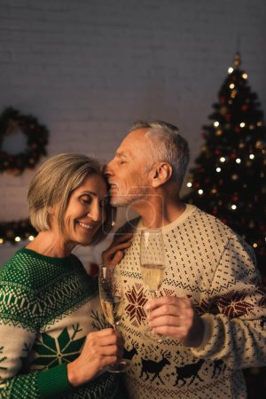 Téléchargez les photos : Homme d'âge moyen en pull festif tenant un verre de champagne et embrassant femme heureuse près de l'arbre de Noël - en image libre de droit