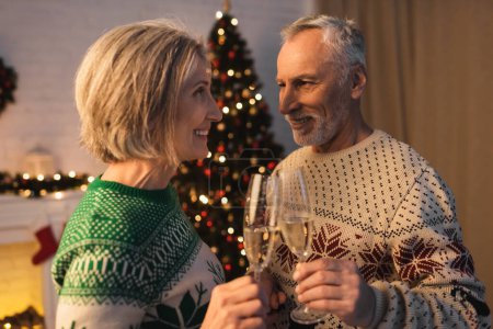 Téléchargez les photos : Joyeux couple d'âge moyen en chandails festifs cliquetis verres de champagne près de l'arbre de Noël flou - en image libre de droit