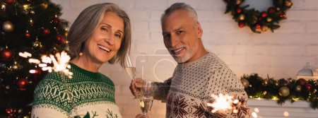 cheerful middle aged couple in festive sweaters holding sparklers and glasses of champagne on christmas eve, banner