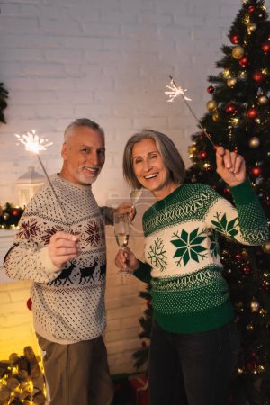 Téléchargez les photos : Joyeux couple d'âge moyen en chandails de fête tenant des étincelles et des verres de champagne la veille de Noël - en image libre de droit