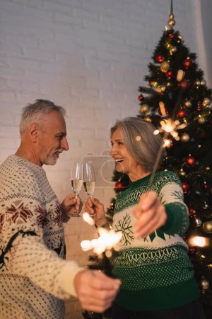 happy mature couple in festive sweaters holding sparklers and glasses of champagne on christmas eve