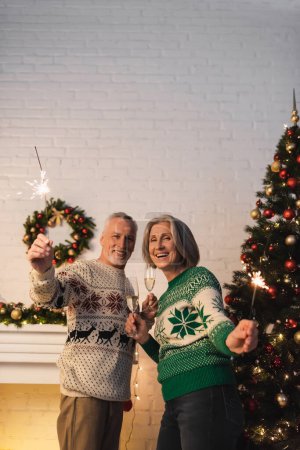 Téléchargez les photos : Heureux couple d'âge mûr dans des pulls tenant des verres de champagne et des étincelles le soir de Noël - en image libre de droit
