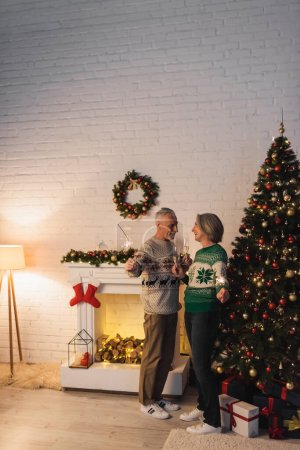 Téléchargez les photos : Heureux couple d'âge mûr dans des chandails tenant des verres de champagne et des étincelles le soir de Noël à la maison - en image libre de droit