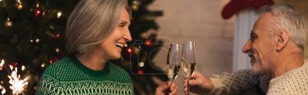 smiling middle aged couple clinking glasses of champagne near shiny sparkler on christmas eve, banner