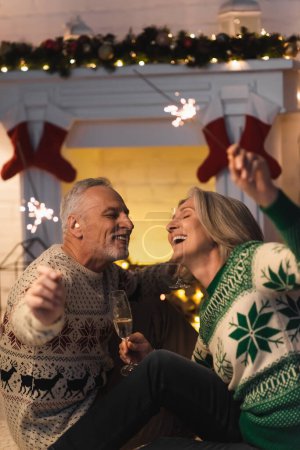 Téléchargez les photos : Joyeux couple d "âge mûr tenant un verre de champagne et des étincelles brillantes la veille de Noël - en image libre de droit