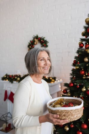 happy and mature woman holding wicker basket near decorated christmas tree at home