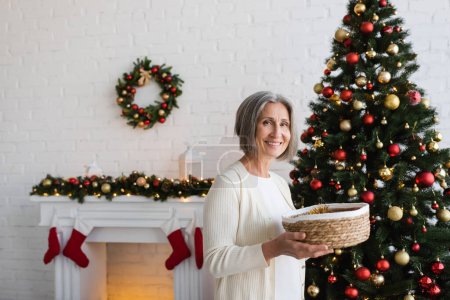 freudige und reife Frau hält Weidenkorb neben geschmücktem Weihnachtsbaum zu Hause