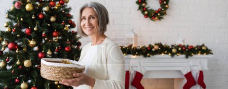 femme gaie et mature tenant panier en osier près décoré arbre de Noël à la maison, bannière