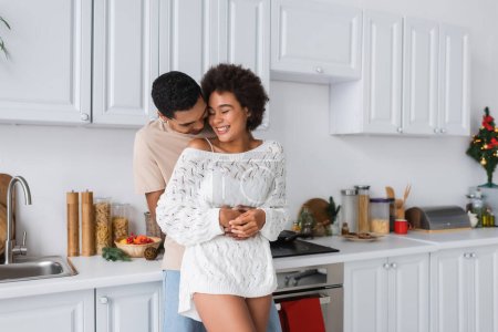 african american man hugging cheerful girlfriend in white knitted sweater in kitchen