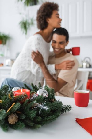 christmas wreath decorated with candle and baubles near blurred african american couple embracing in kitchen