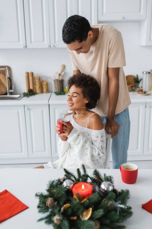 afrikanisch-amerikanische Frau hält rote Tasse in der Hand und lächelt mit geschlossenen Augen neben jungem Freund und dekoriertem Weihnachtskranz