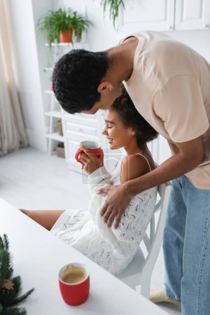 young african american man near smiling girlfriend sitting with cup of cocoa in kitchen