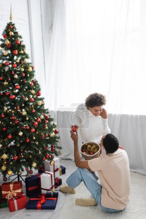 vista de ángulo alto de la pareja afroamericana sosteniendo canasta de mimbre con bolas cerca del árbol de Navidad y cajas de regalo
