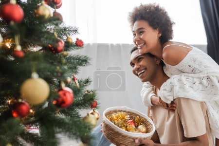 Téléchargez les photos : Joyeux couple afro-américain regardant arbre de Noël décoré près panier en osier avec des boules - en image libre de droit