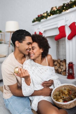 heureux homme afro-américain embrassant petite amie tenant boule de Noël et tinsel dans le panier en osier
