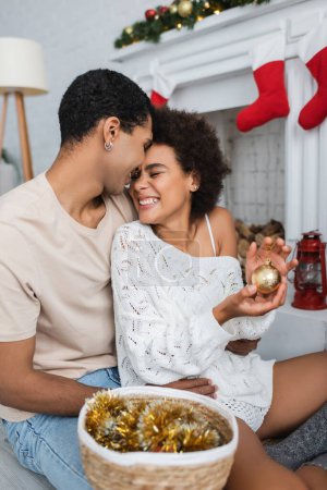aufgeregte Afroamerikanerin hält goldene Weihnachtskugel neben Freund mit Lametta im Weidenkorb