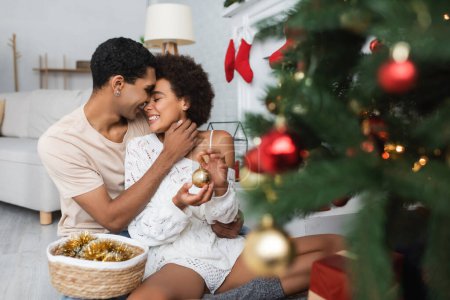 young african american man hugging happy woman with bauble near blurred christmas tree