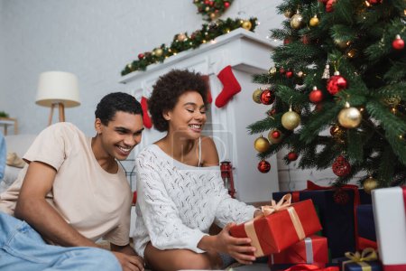 Téléchargez les photos : Heureuse femme afro-américaine en chandail tricoté blanc tenant boîte cadeau près de petit ami et arbre de Noël - en image libre de droit