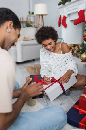 Foto de Feliz africano americano pareja apertura navidad regalos mientras sentado en piso en casa - Imagen libre de derechos