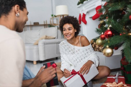 excited african american woman in white openwork sweater sitting with christmas present near blurred boyfriend 