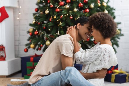 Téléchargez les photos : Vue latérale de jeune et sexy couple afro-américain embrassant près de l'arbre de Noël à la maison - en image libre de droit