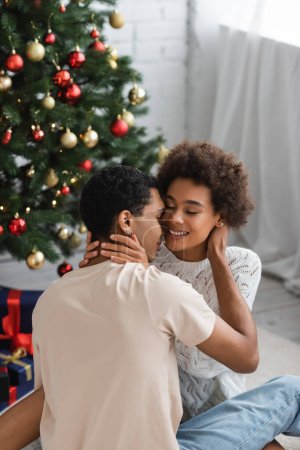Photo for Young african american woman with closed eyes hugging boyfriend on christmas day at home - Royalty Free Image