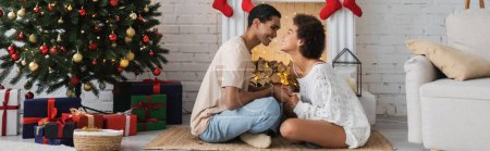 Téléchargez les photos : Vue latérale de heureux couple afro-américain tenant la main sur le sol près de l'arbre de Noël et la cheminée, bannière - en image libre de droit
