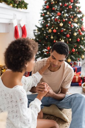 Photo for Young african american man holding hands of girlfriend touching his neck near blurred christmas tree - Royalty Free Image