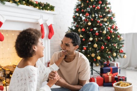 Photo for Cheerful african american couple holding hands and looking at each other near christmas tree and fireplace - Royalty Free Image