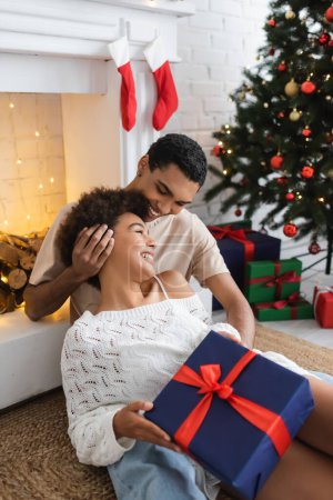lächelnder afrikanisch-amerikanischer Mann am Kamin sitzend mit glücklicher Freundin, die ein Weihnachtsgeschenk hält
