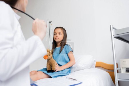Téléchargez les photos : Fille tenant jouet doux près flou médecin et presse-papiers dans la salle d'hôpital - en image libre de droit