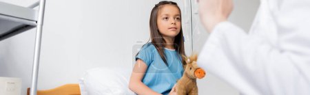 Niño con juguete suave mirando al pediatra borroso en la sala del hospital, pancarta 