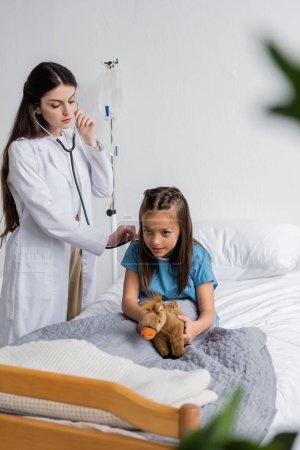 Doctor checking lungs of child with stethoscope in hospital ward 