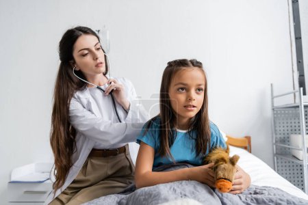 Kid with soft toy sitting near doctor with stethoscope on hospital bed 