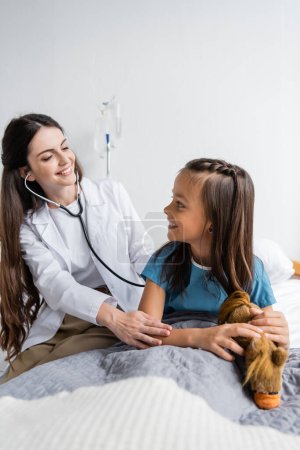 Cheerful pediatrician with stethoscope calming patient near soft toy on hospital bed 