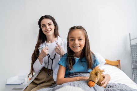 Smiling kid holding soft toy near doctor with stethoscope looking at camera in hospital ward 