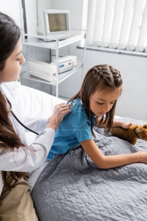 Pediatrician with stethoscope checking back of patient on hospital bed  
