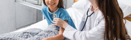 Cropped view of pediatrician holding stethoscope near chest of kid on bed in clinic, banner 