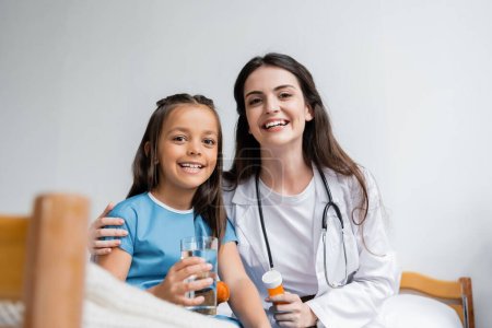 Foto de Pediatra sonriente y niño con bata de paciente sosteniendo un vaso de agua y pastillas en la sala del hospital - Imagen libre de derechos