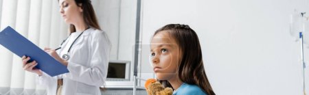 Photo for Kid with toy looking away near blurred pediatrician holding clipboard in clinic, banner - Royalty Free Image